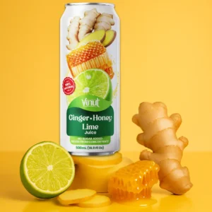 A can of Vinut Ginger-Honey Lime Juice is displayed on a yellow surface with fresh ginger, a halved lime, and honeycomb in front.